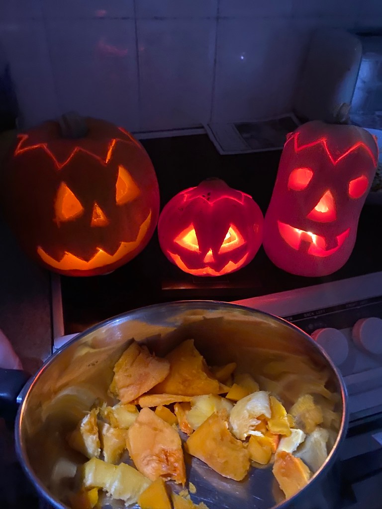 Three carved pumpkins with illuminated faces, displaying Halloween themes, sit on a countertop, with a pot of pumpkin innards in the foreground.